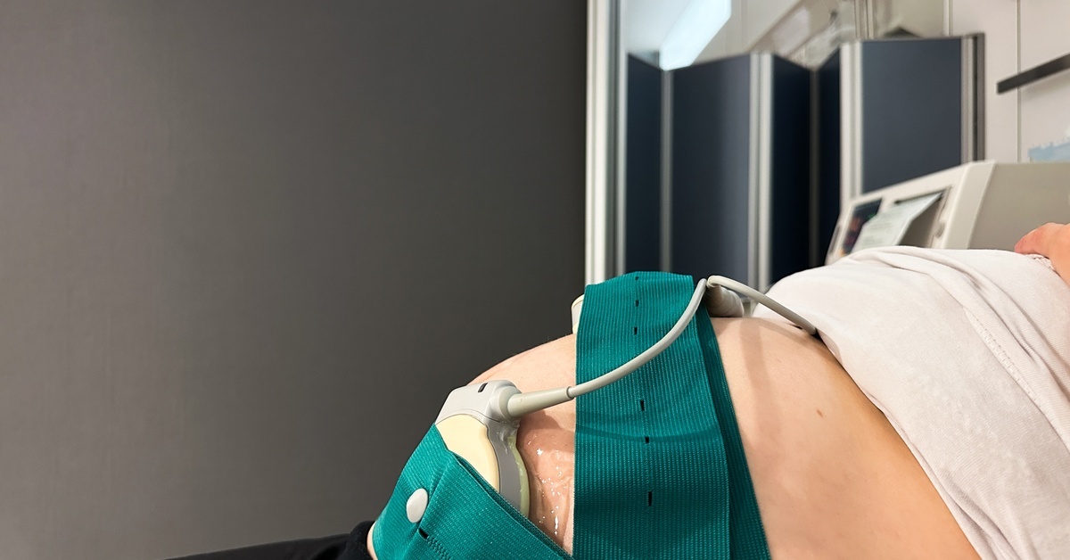 A pregnant woman lies on a medical table. Green bands are wrapped around her exposed belly, holding a fetal monitor in place.