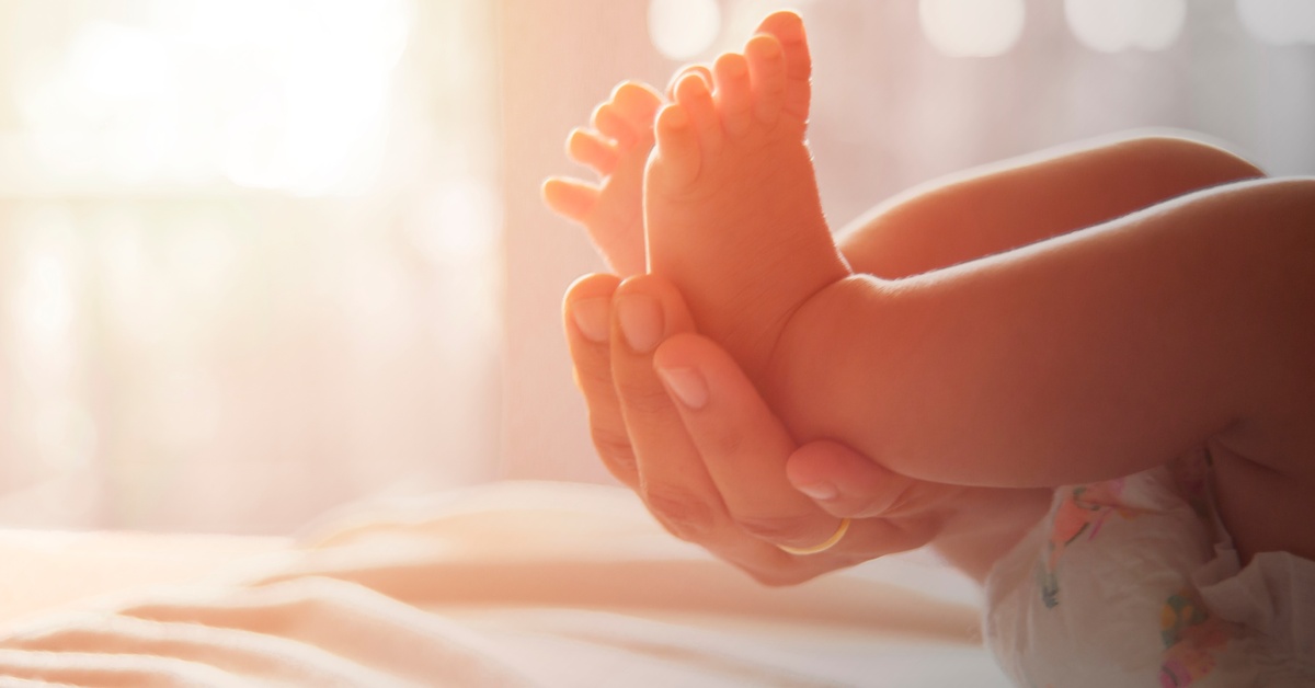 A close-up view shows the feet of a newborn baby being held up by a hand. The sunshine illuminates the baby's feet.