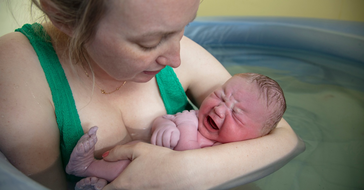 A close-up view shows a mother sitting in a home birthing pool filled with water. She's nestling her baby in her arms.