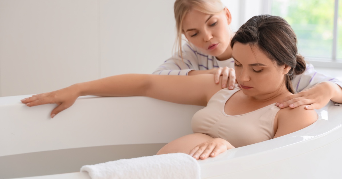 A young woman sits in a white bathtub with her eyes closed. A woman appears behind her resting her hands on her shoulders.