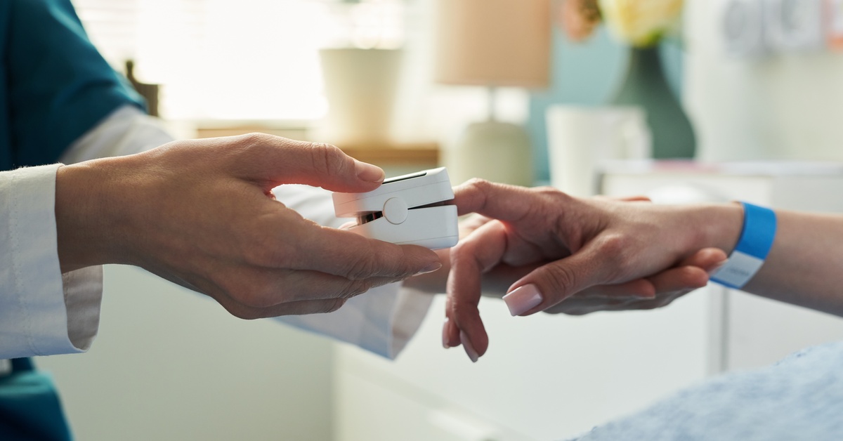A medical professional is placing a pulse oximeter on a patient who's lying in a hospital bed. The background is blurry.
