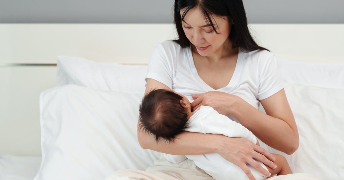 A woman wearing a white T-shirt sits in a bed with white sheets breastfeeding an infant in a white onesie.