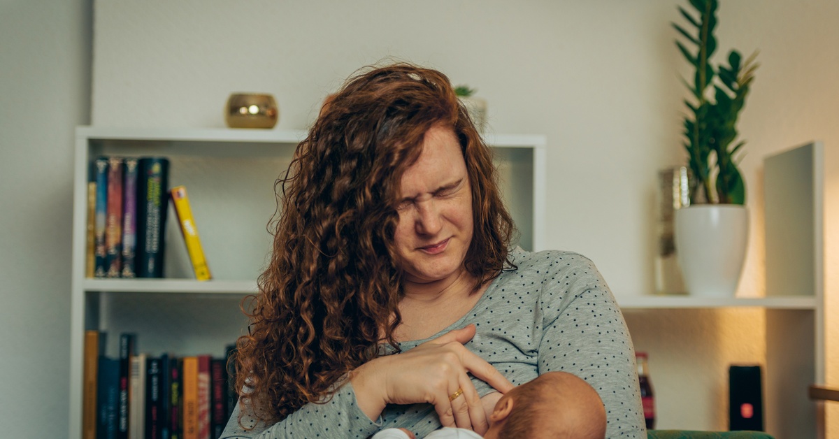 A woman wearing a gray polka dot shirt is breastfeeding an infant. She has her eyes closed and a painful look on her face.