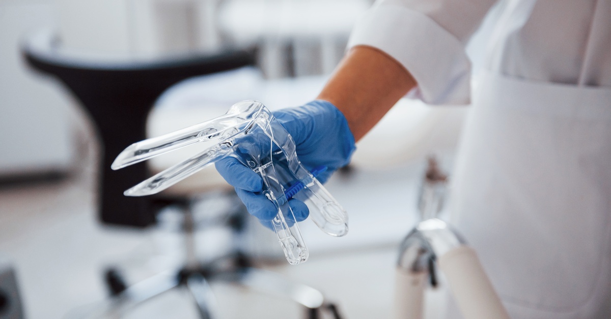 A medical professional wearing a white lab coat holds a clear, plastic speculum. The background is blurry.