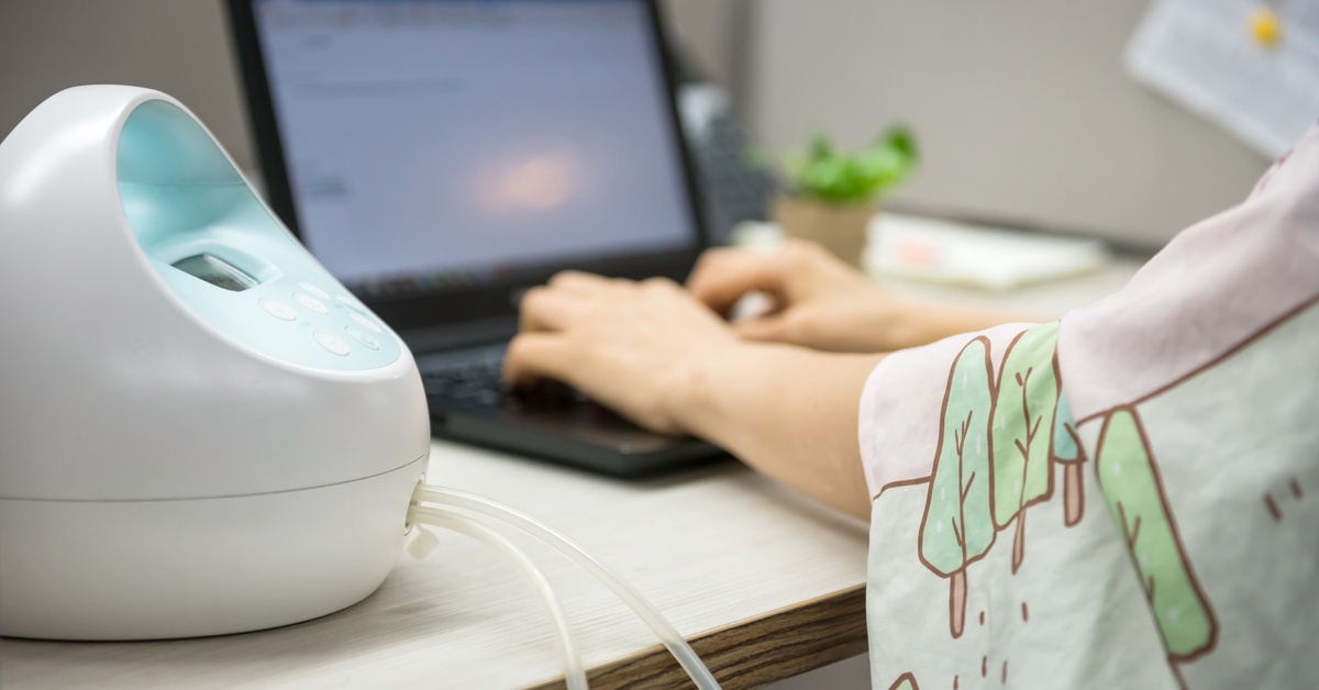 A close-up view shows a white and teal breast pump motor sitting on a table next to a person typing on a laptop.