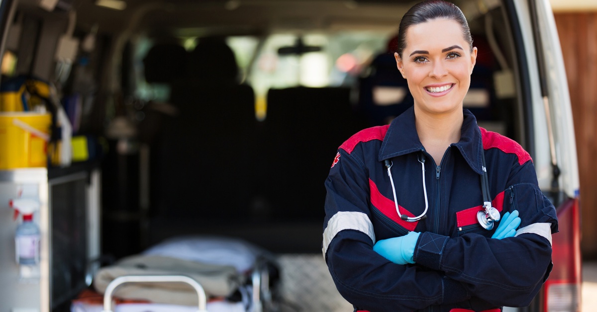 A female EMT stands in front of the back of an emergency service vehicle that has its doors open.