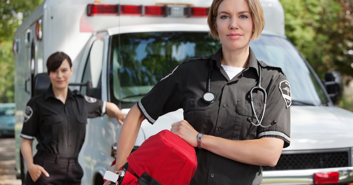 Two female EMTs appear by an ambulance. One is holding a red bad and one is leaning on the ambulance.