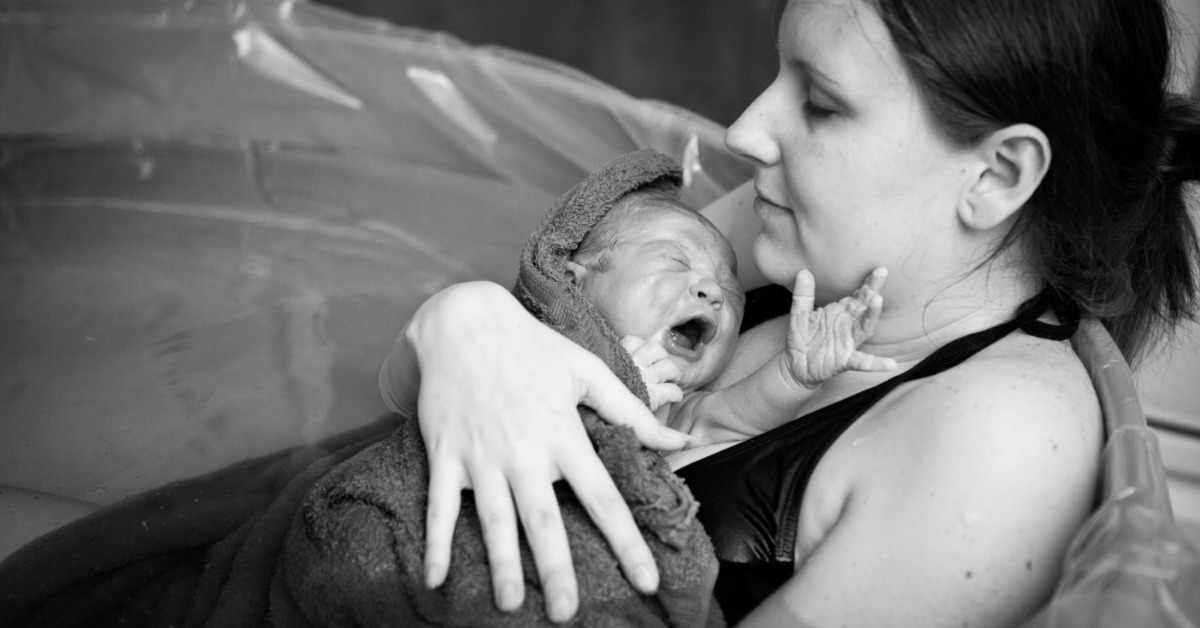 A black and white image shows a mother holding her newborn baby wrapped in a towel after giving birth in a tub.
