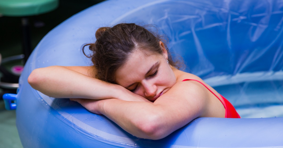 A woman wearing a bright red spaghetti strap top rests her arms and head on the side of a birthing pool.