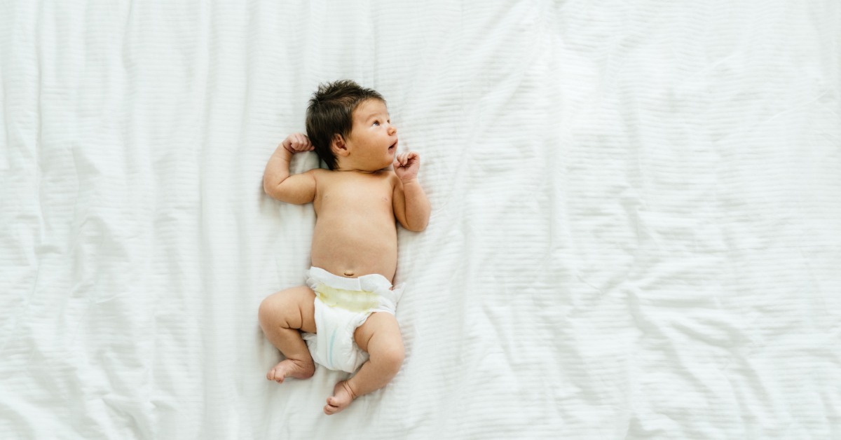 An aerial view shows an adorable newborn baby with lots of black hair lying on their baby on a white blanket.
