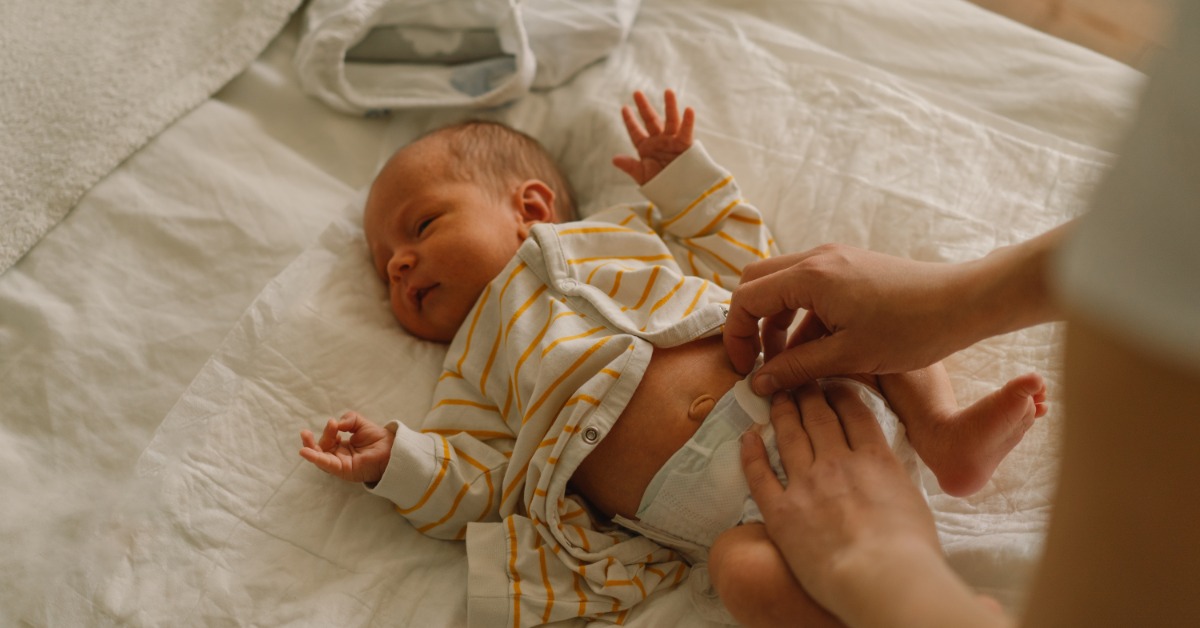 A newborn baby is lying on a white blanket while a person is beginning to remove the tabs on the baby's diaper.