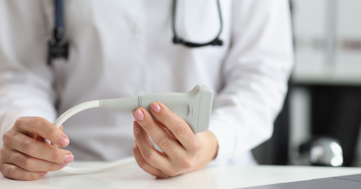 A close-up view shows a medical professional resting their hands on a white surface while holding a gray Doppler probe.