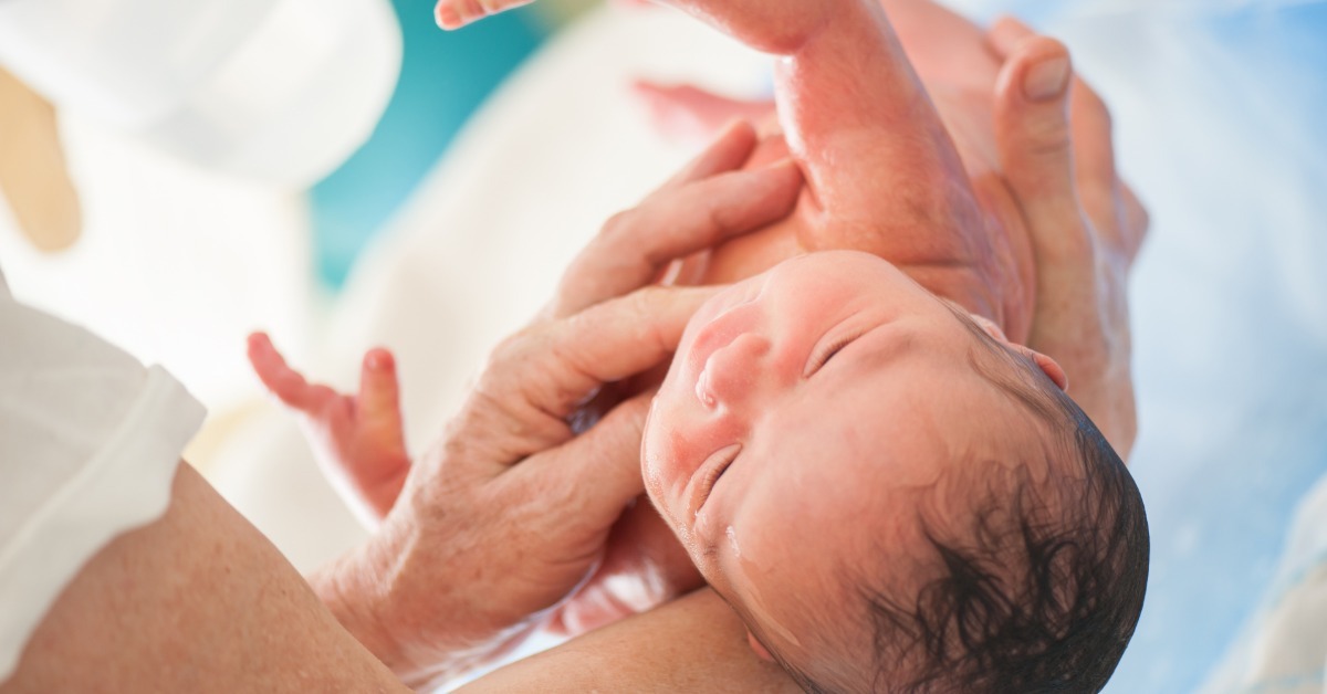 A close-up view shows a newborn baby being held above water by an adult. The baby's skin is wet.