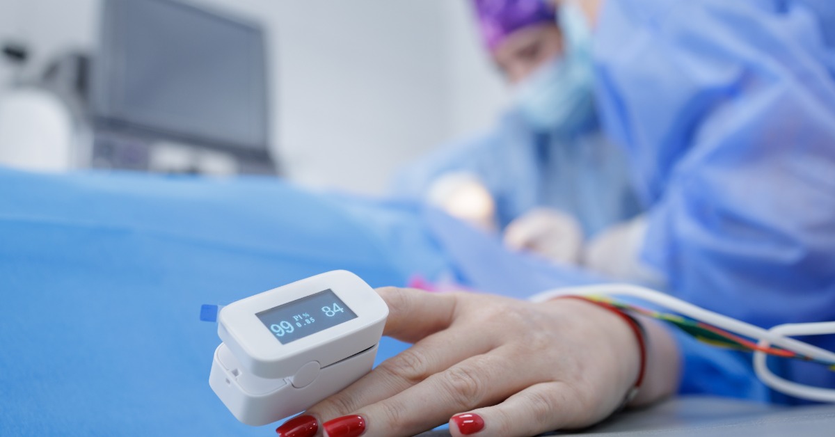 A close-up view shows a pulse oximeter on the hand of a patient lying on a medical table. Doctors are behind them.