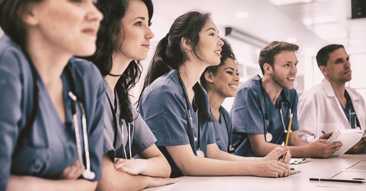 Four female and two male healthcare professionals sit and lean on a table. They are actively listening and learning.