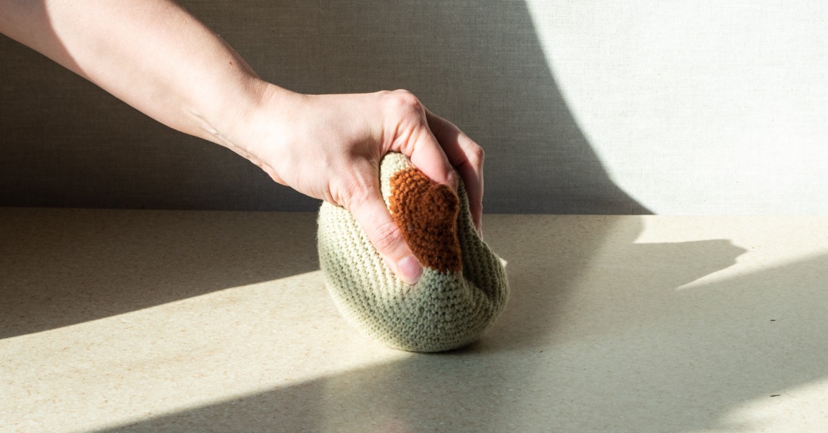 A close-up view shows a person's hand squeezing a knitted breast model. The sunshine creates a shadow.