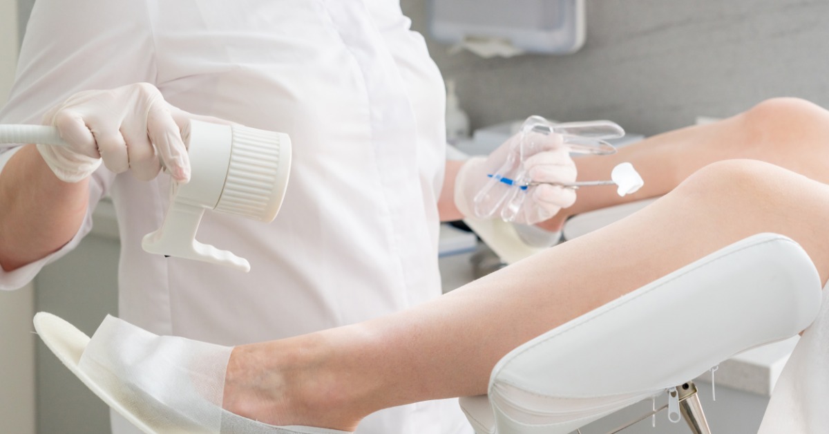 A medical professional holds a speculum in one hand while adjusting a light with the other. A patient lies on an exam table.