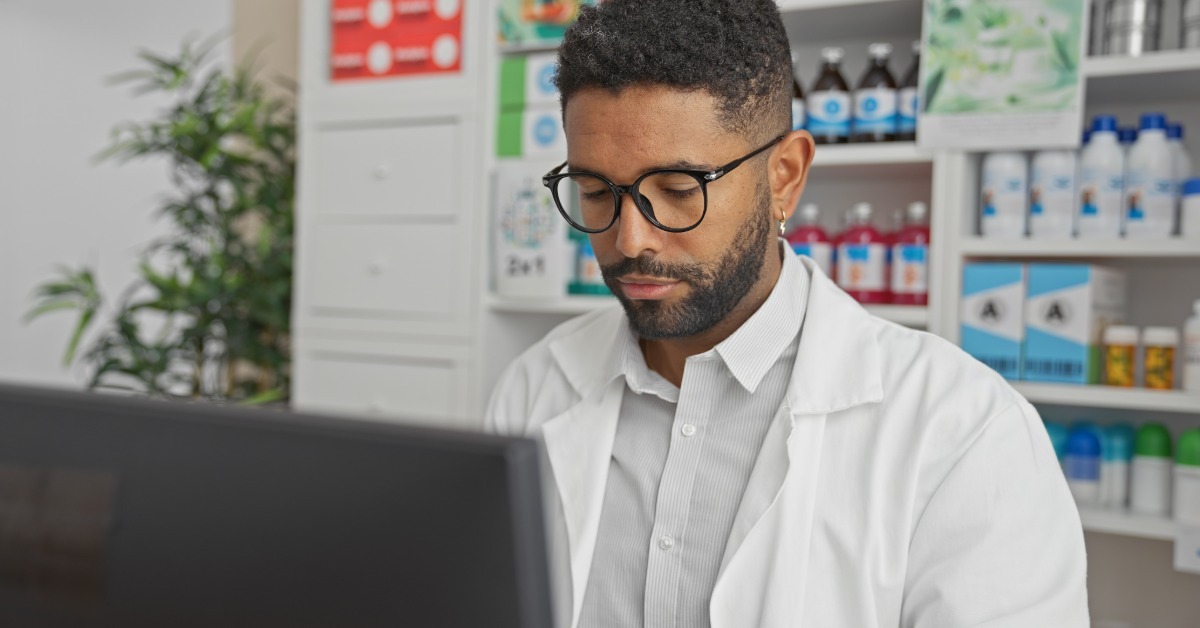 A male healthcare professional is behind a work computer, looking down. Medical supplies are behind him.