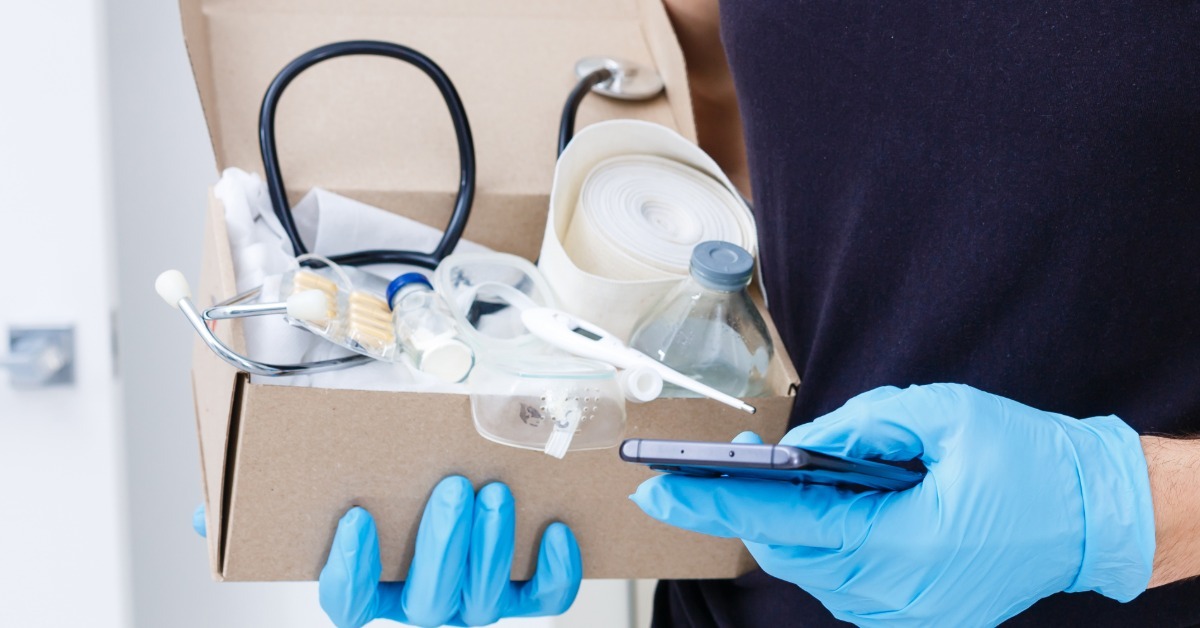 A close-up view shows a person wearing blue latex gloves holding a phone in one hand and a box of medical supplies in the other.