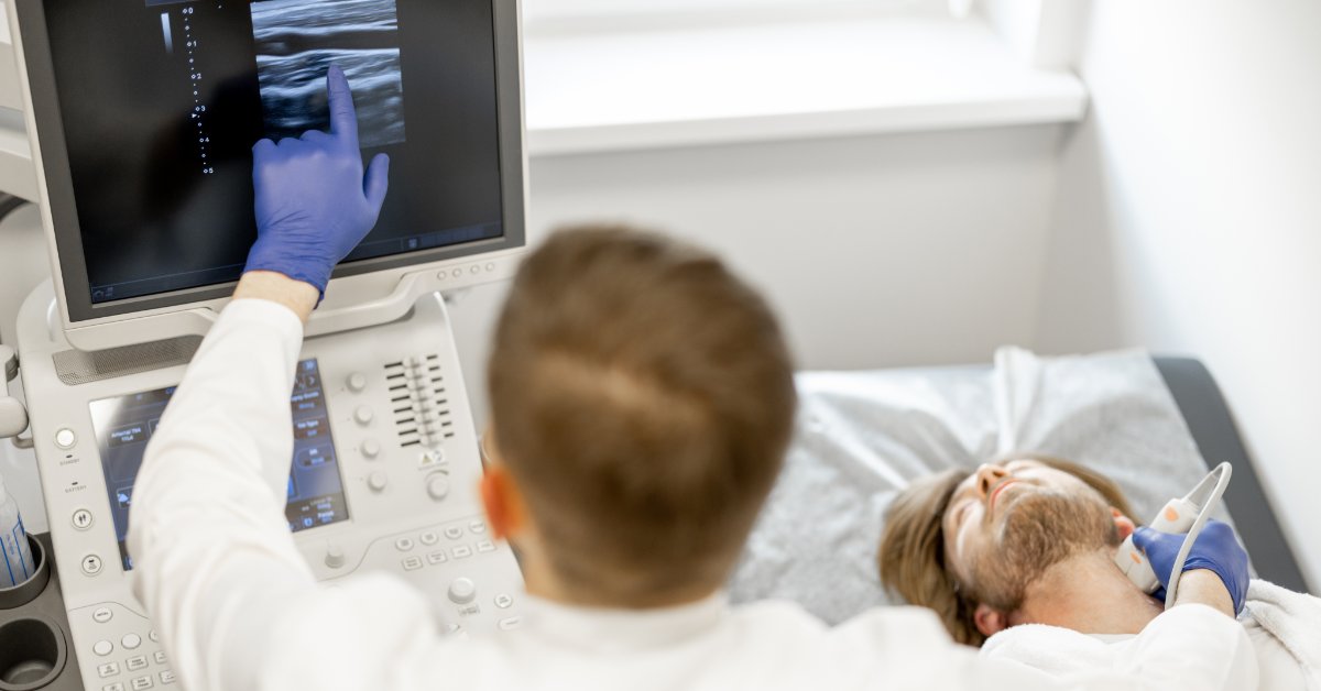 A medical professional is pressing a Doppler on the neck of a male patient and pointing to the ultrasound screen.