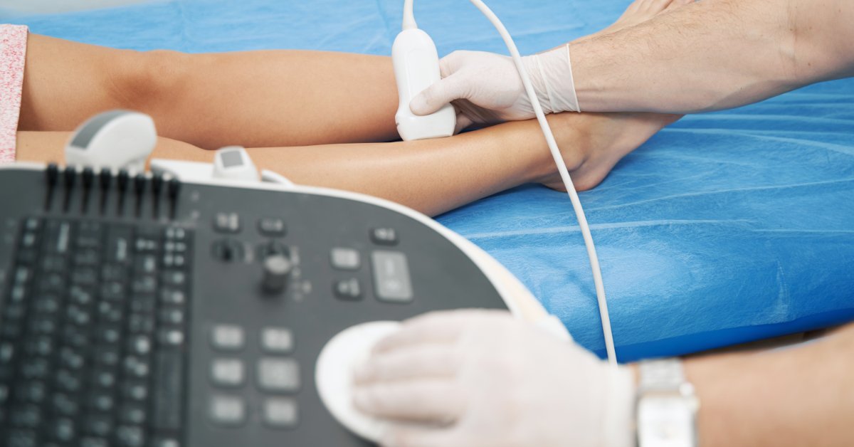 A medical professional wearing white latex gloves is pressing a Doppler onto the lower part of the leg of a patient.