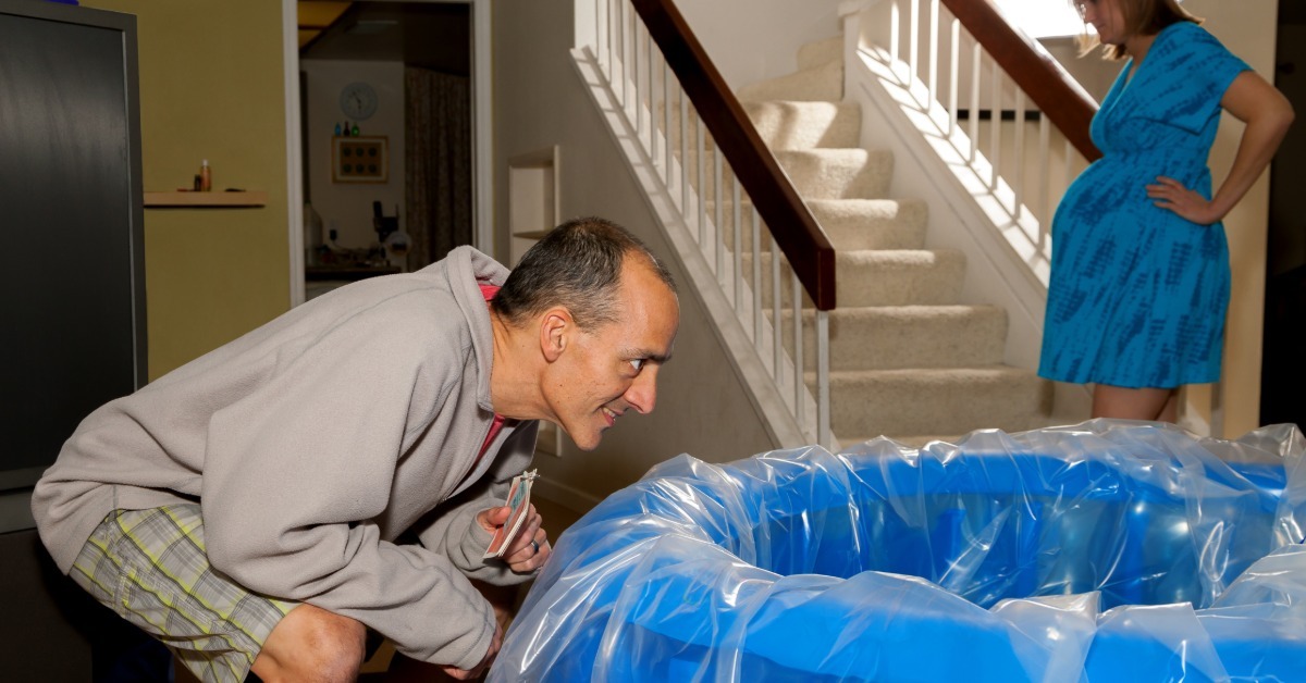 A man is squatting to inspect a blue birthing pool while a pregnant woman leans against the staircase railing.