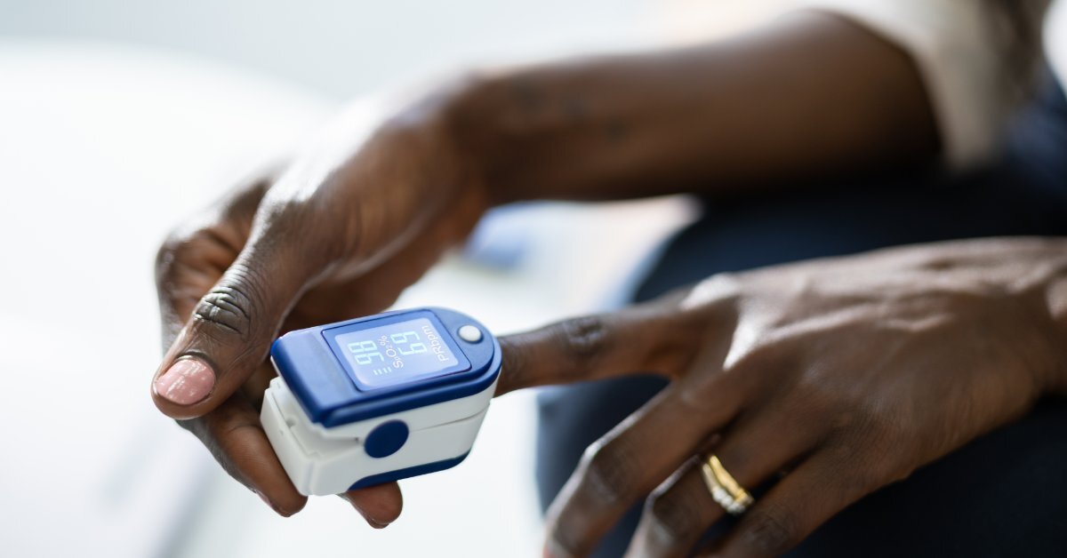 A close-up view shows a person placing a white and blue pulse oximeter onto their left index finger.