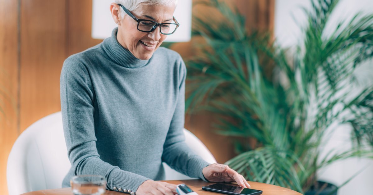 A smiling woman sits at small wooden table looking at the pulse oximeter that sitting on her right index finger.
