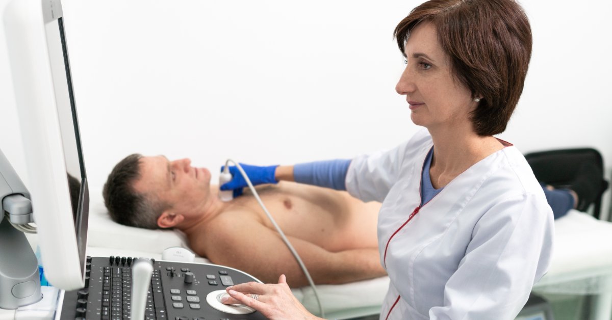 A female medical professional places a Doppler probe on a male patient's neck while looking at the Doppler monitor.