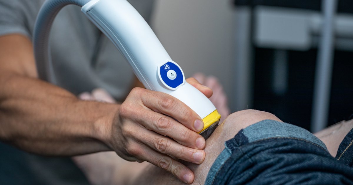 A close-up view shows a white, blue, and yellow Doppler probe that a medical professional is placing on a patient's knee.