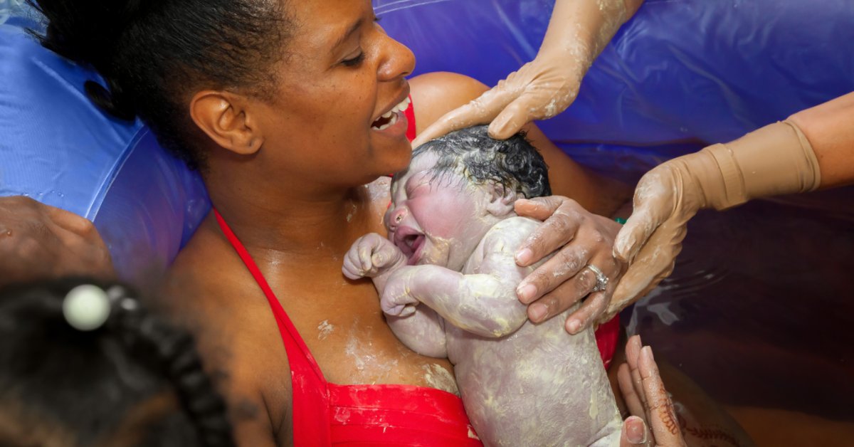 A close-up view shows a smiling woman wearing a red bathing suit top holding a newborn baby while sitting in a birthing pool.