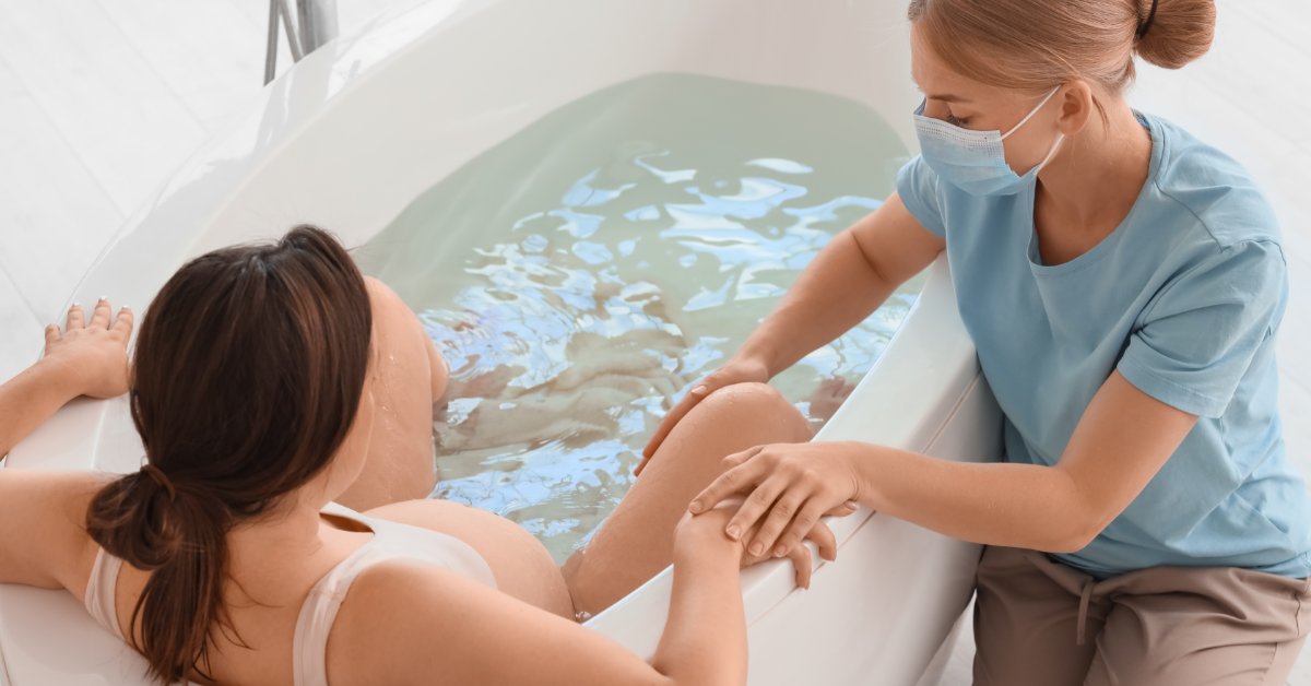 A pregnant woman sits in a white bathtub filled with water. A medical professional sits on the side holding her hand and leg.