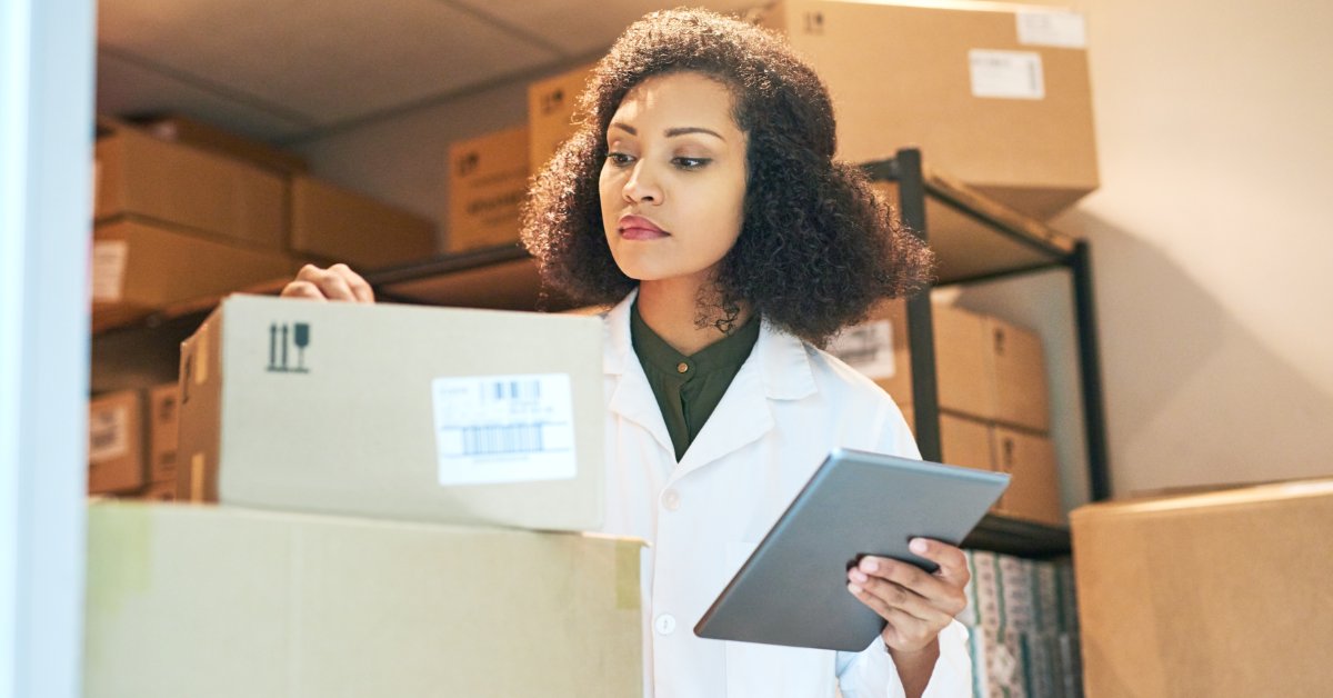 A female medical professional holds a tablet while looking a small cardboard box that sits on top of other boxes.