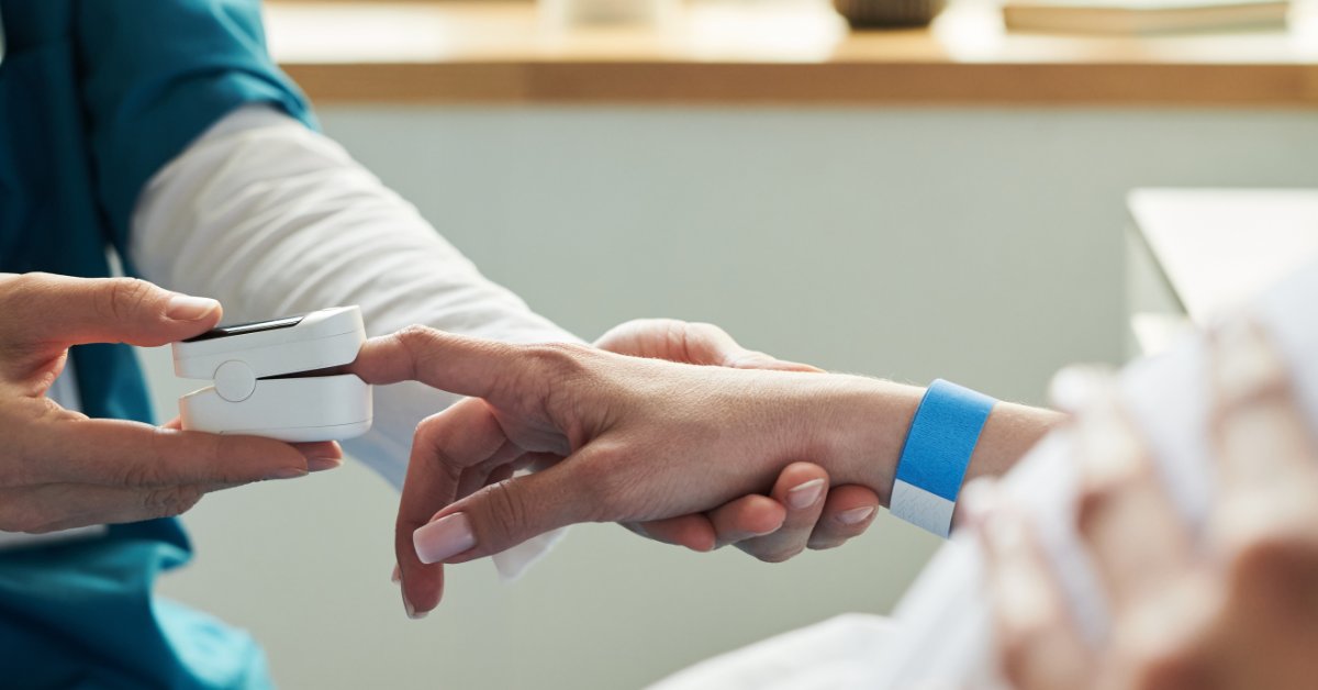 A medical professional is holding a patient's hand and placing a white pulse oximeter on their index finger.