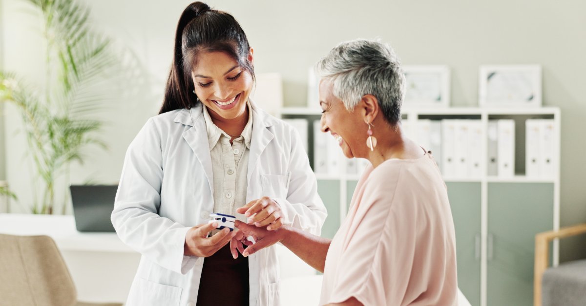 A female medical professional is smiling and looking at a pulse oximeter that's on the smiling female patient's finger.