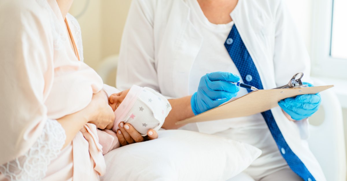 A woman wearing a pink bathrobe is sitting down next to a medical professional breastfeeding a baby.
