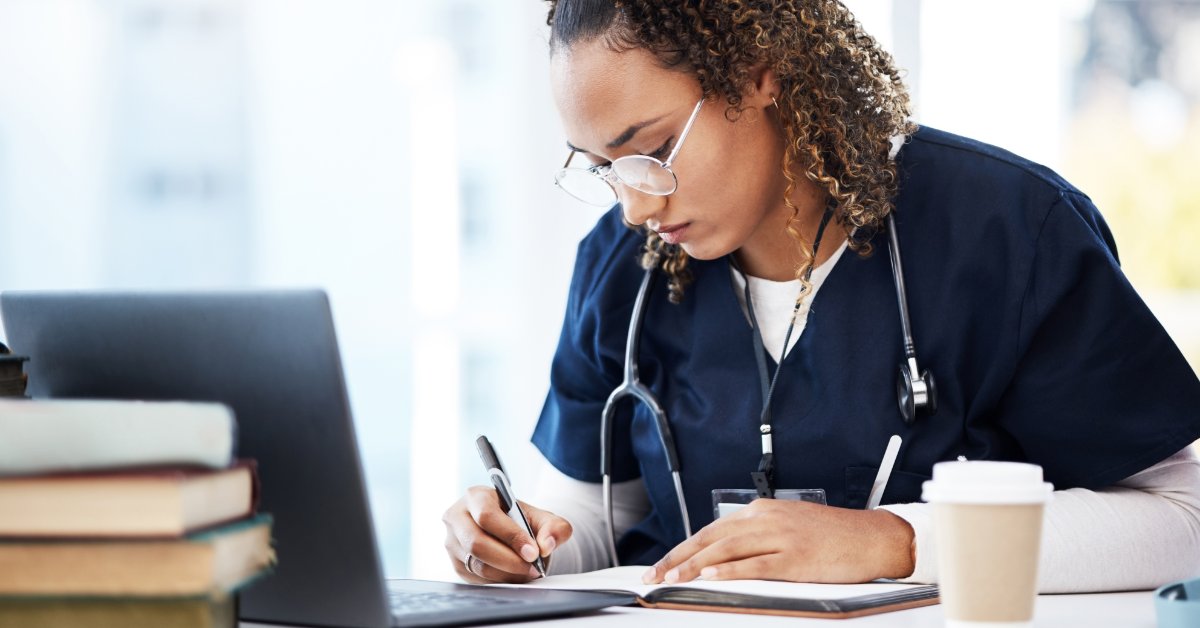 A female medical professional sits at a desk writing in a notepad. A coffee cup, a laptop, and books also appear on the desk.