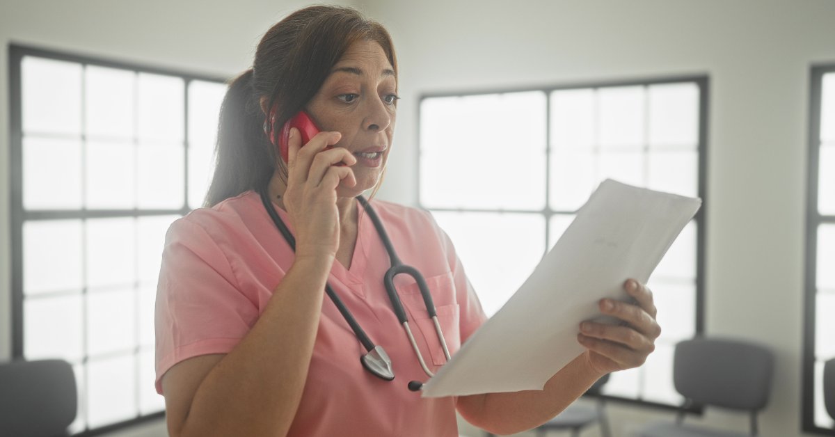 A medical professional wearing pink scrubs holds a red phone up to her ear and is looking at a piece of paper.