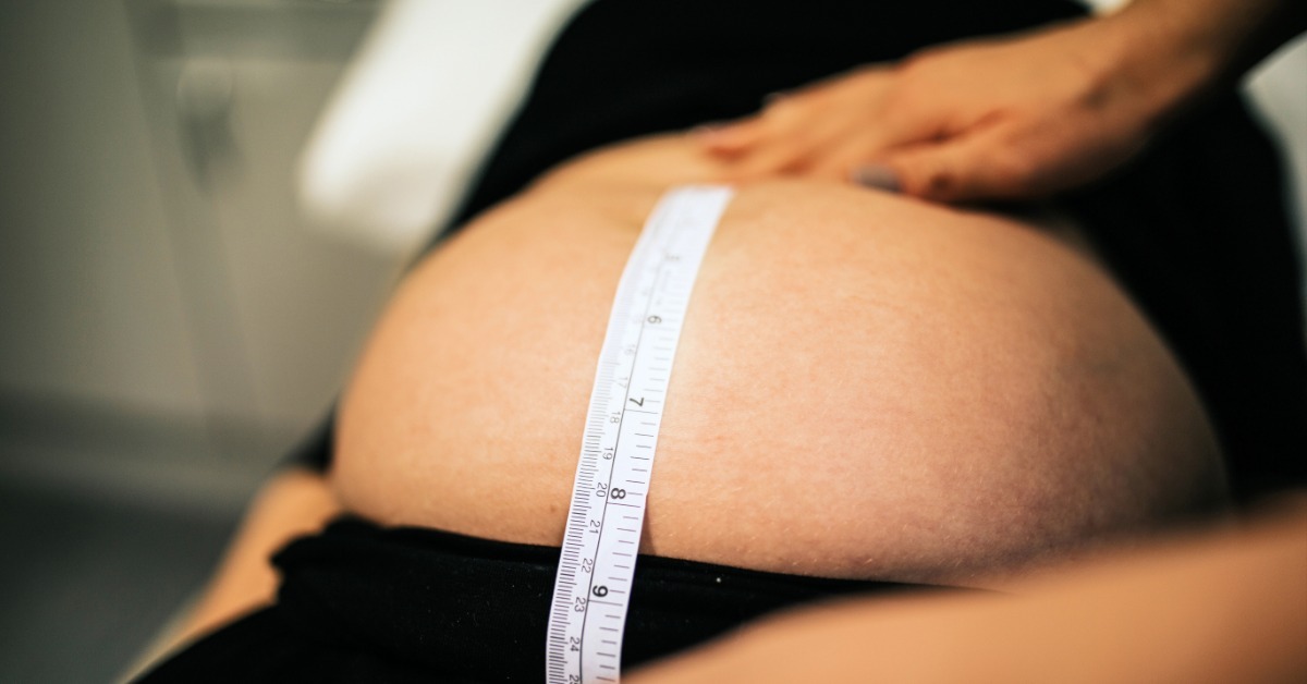 A close-up view shows a medical professional using a flexible tape measure to measure a pregnant woman's belly.