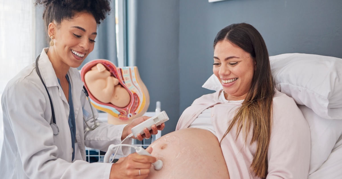A smiling healthcare professional holds the probe of a fetal Doppler to the bare stomach of a smiling pregnant woman.