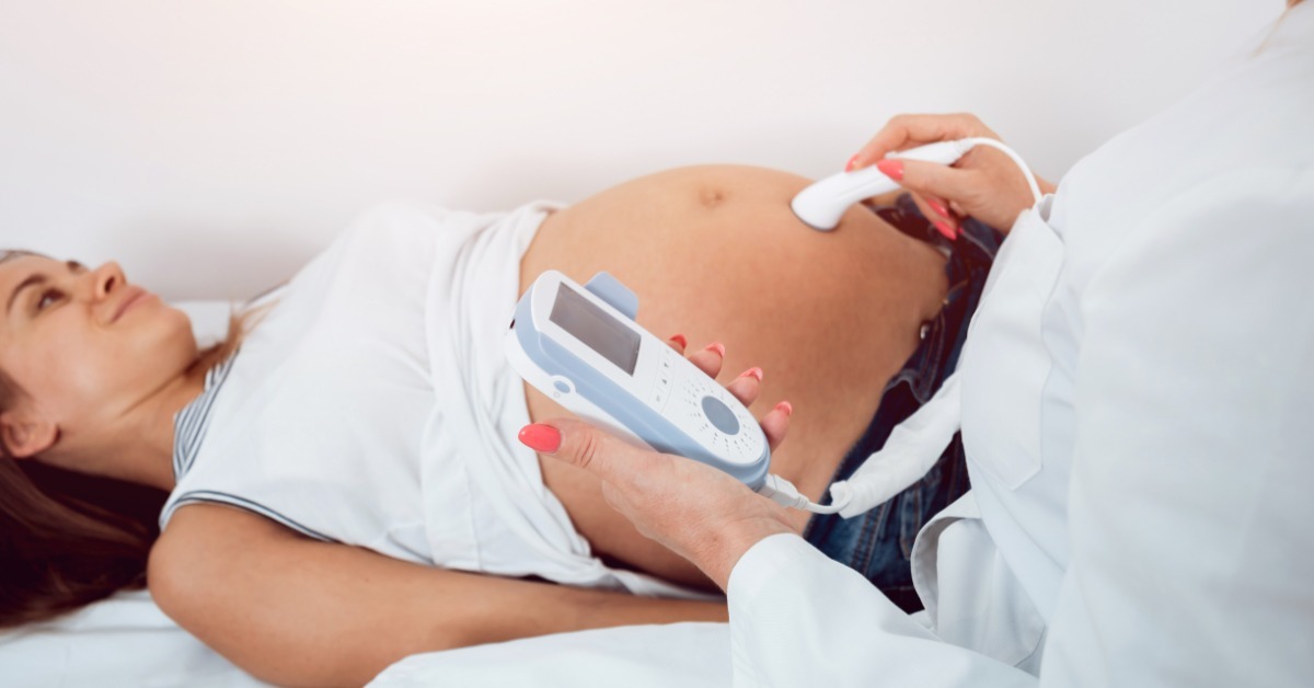A pregnant female patient lies on an exam table while a healthcare professional uses a fetal Doppler probe on her stomach.