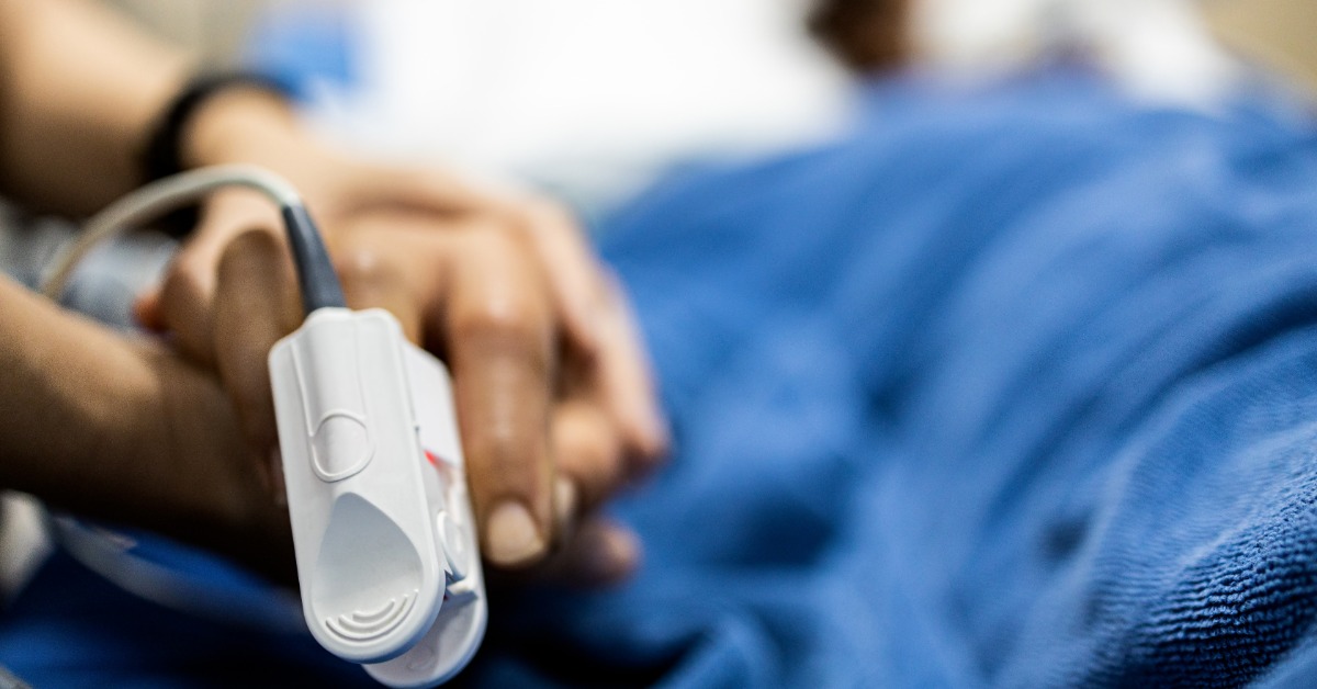 A close-up view shows a pulse oximeter attached to the middle finger of a patient who is lying in a hospital bed.