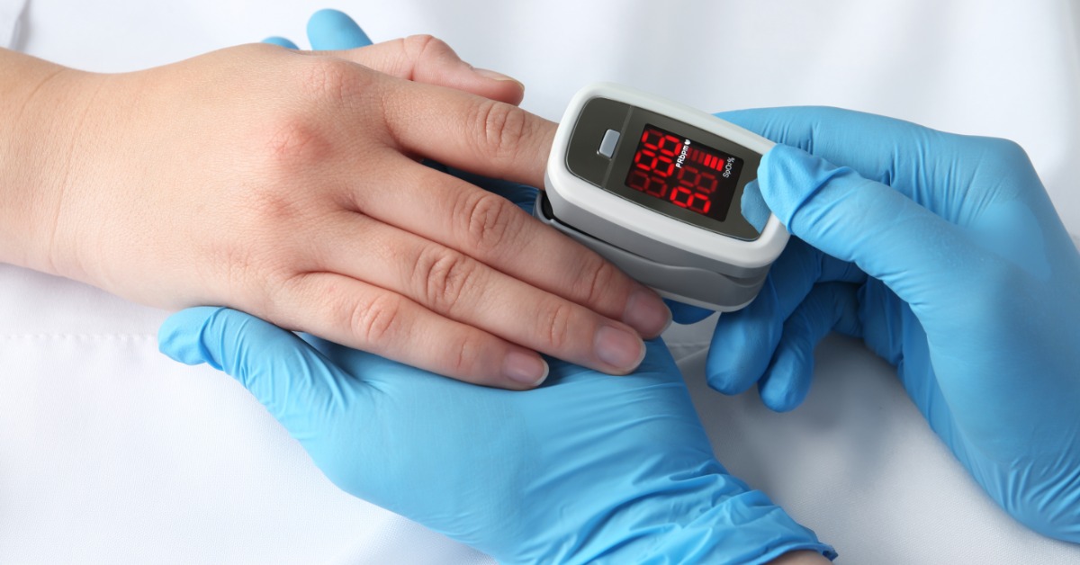 A healthcare professional wearing blue latex gloves is putting a pulse oximeter on the pointer finger of a patient.