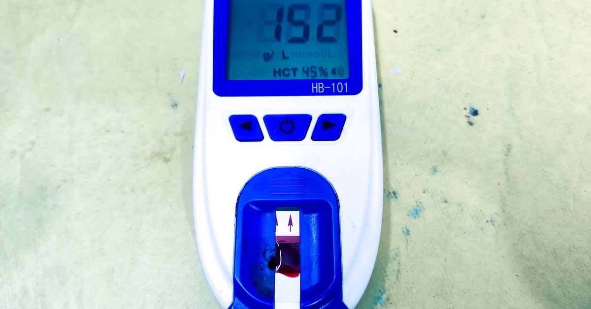 A digital hemoglobin analyzer displays a reading of 15.2 g/L with HCT 45% on the screen, with a test strip inserted at the bottom.