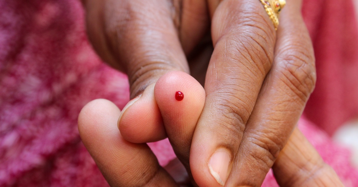 A hand holds a finger with a small drop of blood on the fingertip, showing the result of a finger prick for hemoglobin testing.