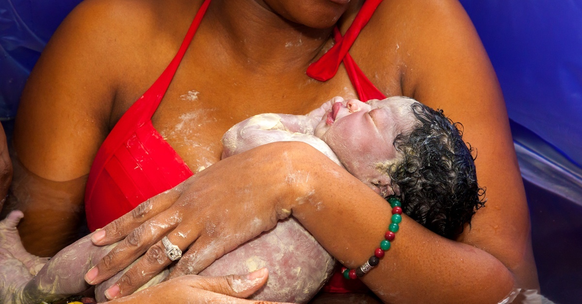 A birthing parent holds a newborn in an inflatable birth pool. Vernix caseosa covers the baby after the water birth.