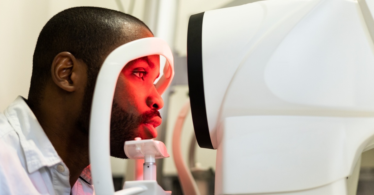 A dark skinned man rests his head on the equipment for a cornea exam. The equipment illuminates his face in red.