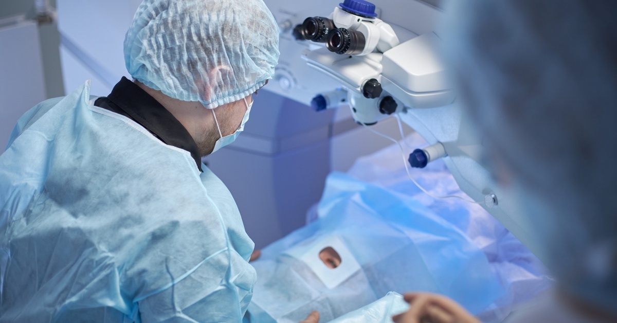 A surgeon sits over a patient who is resting on an exam table. There is surgical draping covering her face except for one eye.