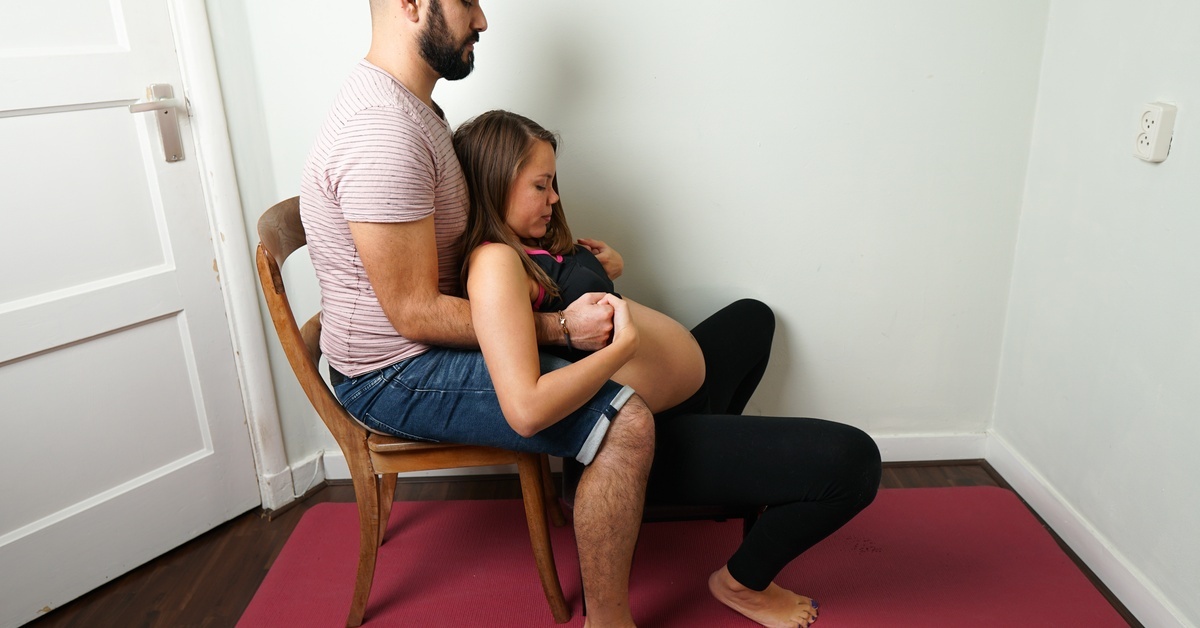 Positions for Out-of-Bed Laboring During a Home Birth A man and pregnant woman rehearse a birth position. The man sits in a chair and holds the woman by her torso off the ground.